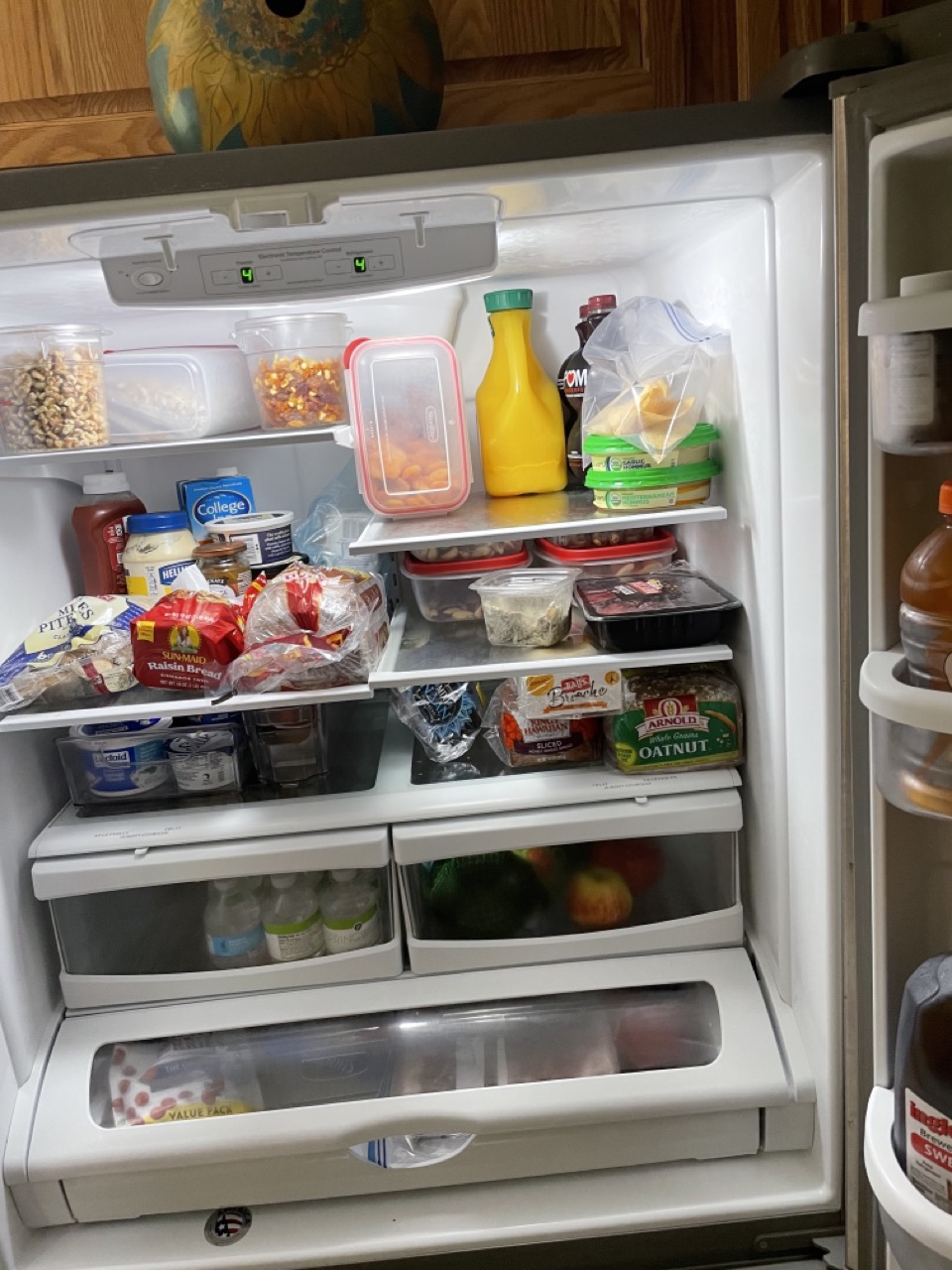 The Haunted Bowling Alley: The picture shows an open refrigerator filled with various food items. On the top shelf, there are containers with nuts and snacks, a bottle of orange juice, and some other bottles and containers. The second shelf has a mix of items including bread, butter, and a plastic container. The third shelf has a pack of sliced cheese, a loaf of bread, and some other items. Below that, there are two crisper drawers with fruits and vegetables visible through the transparent front. The inside of the refrigerator door has several shelves with condiments and bottles. Above the fridge, there is a painting or picture of a bird. The refrigerator is white and has digital temperature controls on the top.