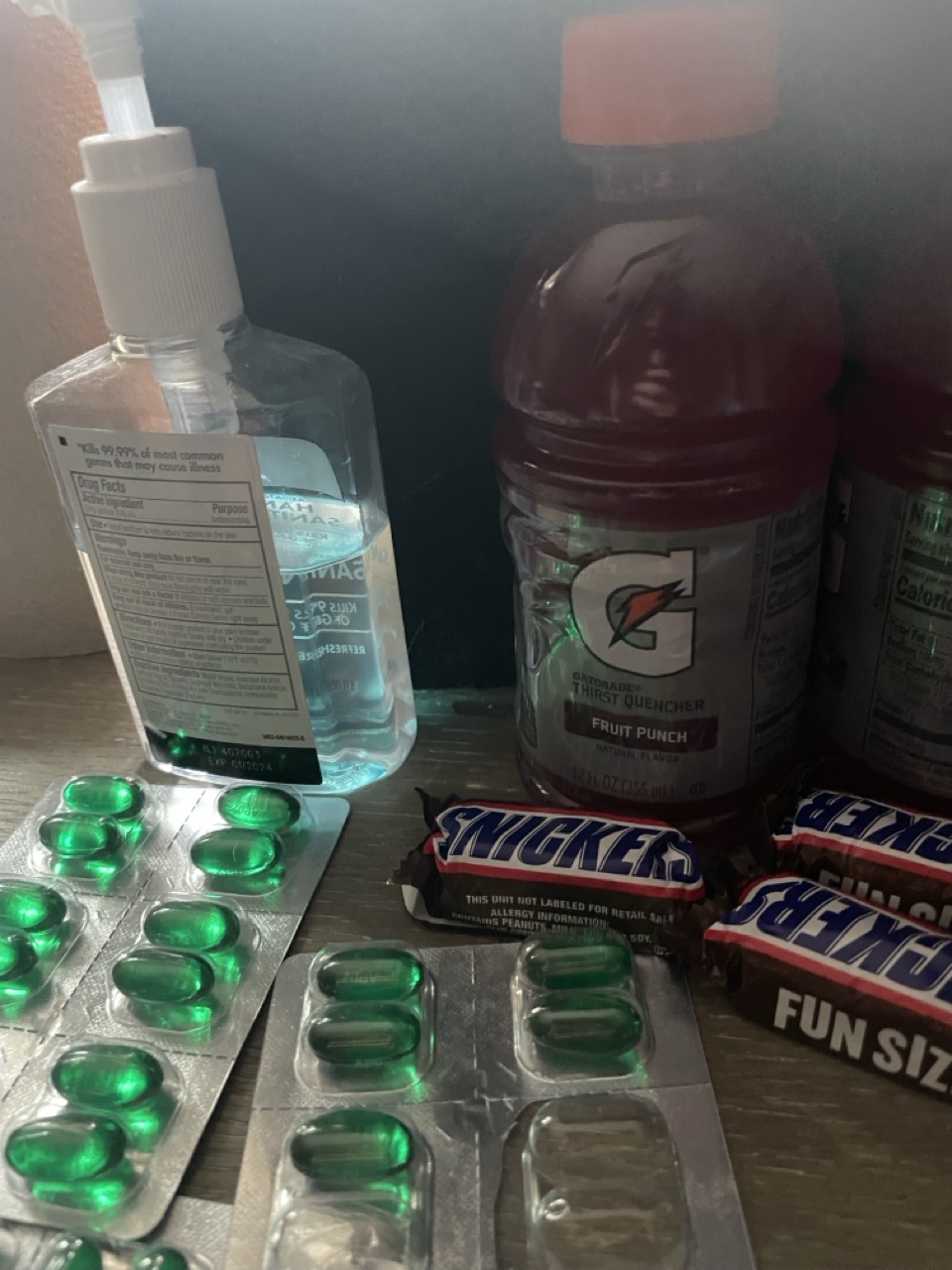 The Covid Sucker Punch: The picture shows a few items on a surface. On the left, there is a clear bottle of hand sanitizer with a white pump. Next to it, there is a red Gatorade bottle with a label that says "Thirst Quencher Fruit Punch". Below these items, there are three blister packs of green gel capsules. On the right side, there is a Snickers chocolate bar with a blue and white wrapper. The background is dark and the surface appears to be wooden.