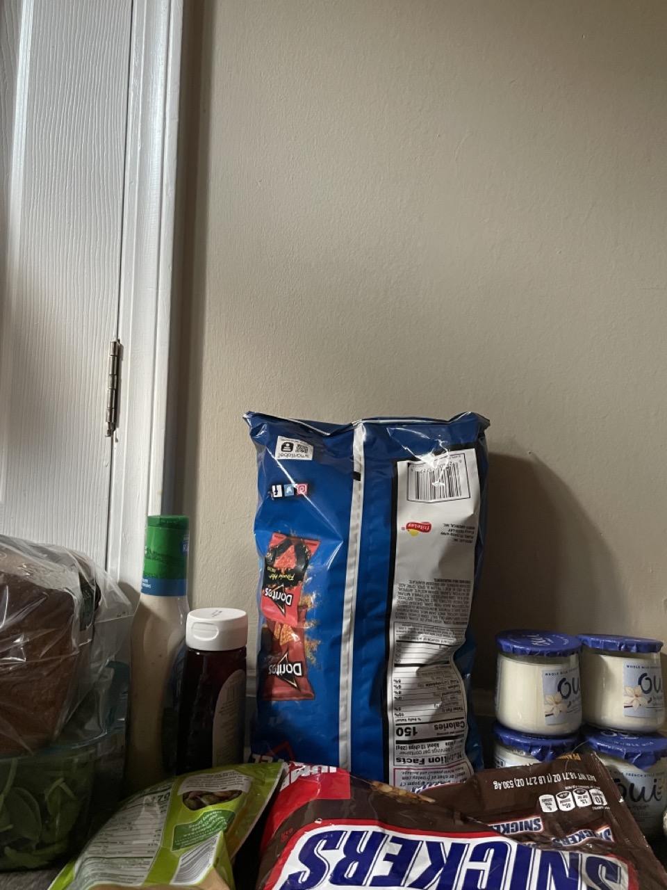 The Timeless Dinner Club: The picture shows a corner of a room with a variety of food items. On the left side, there is a white door frame and a beige wall. The food items are placed on the floor against the wall. There is a large blue bag of Doritos chips leaning against the wall. In front of it, there are several smaller food items including a green bag of snacks, a red bag of Snickers mini candy bars, and three containers of Oui yogurt. There is also a bottle with a green cap that looks like a sauce or dressing, and a brown bottle next to it. The items are arranged haphazardly.