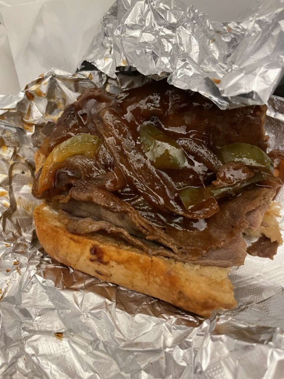 The Not Really Cheesesteak: The picture shows a close-up of a sandwich that is partially wrapped in aluminum foil. The sandwich appears to have roast beef with a rich, dark brown sauce on it. There are also some sliced mushrooms and onions in the sandwich. The bread looks toasted and has a golden-brown color. The aluminum foil is crinkled and reflects light. It seems like the sandwich is freshly made and ready to be eaten.