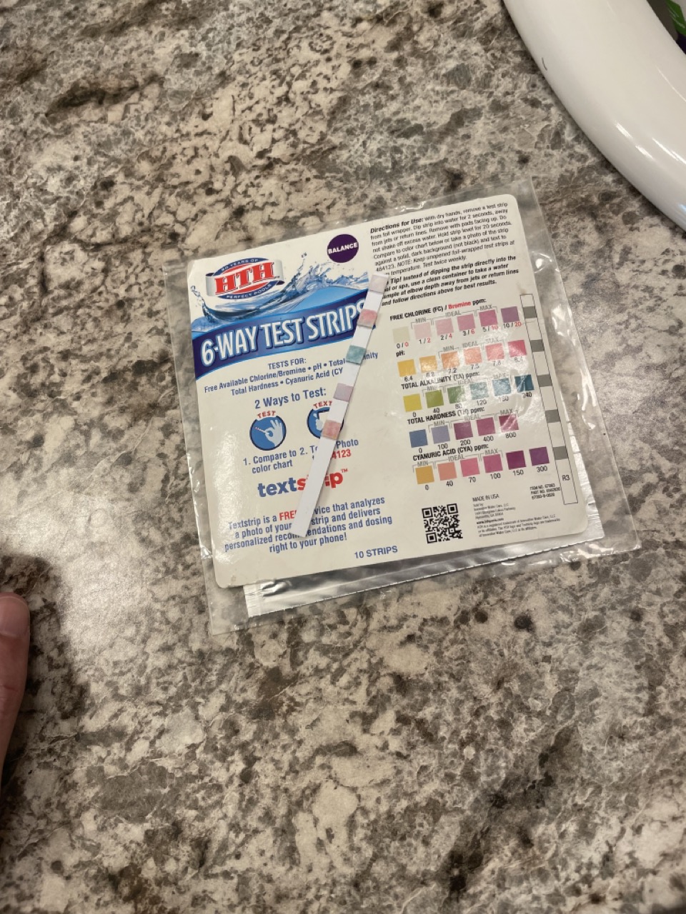 The Adventure of Perfect Balance: The picture shows a pack of HTH 6-Way Test Strips lying on a marble countertop. The pack is labeled "6-Way Test Strips" and has a blue wave design at the top. It mentions that it tests for Free Available Chlorine/Bromine, pH, Total Alkalinity, Total Hardness, and Cyanuric Acid. There are instructions on the pack that mention two ways to test: 1. Compare strip to color chart on the back of the bottle. 2. Text a photo of your strip and get a personalized recommendation and dosing right to your phone. The pack also has a color chart with different colors and values for pH, Total Alkalinity, Total Hardness, and Free Available Chlorine/Bromine. There is also a QR code and it mentions that the pack contains 10 strips.