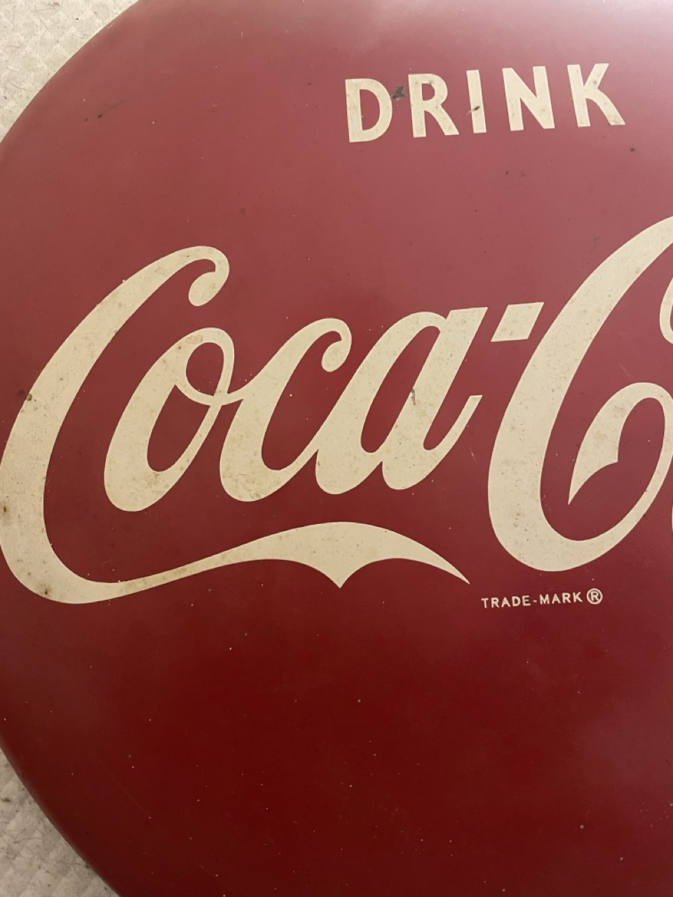 The Mob of GMO Clones: The picture is of a vintage Coca-Cola sign. The background is a deep red color. The word "Drink" is written in capital letters at the top in a cream color. Below it, there is the iconic Coca-Cola logo in cursive, also in cream color. The sign has a round shape and shows some signs of wear and tear, giving it an antique look. At the bottom right, there is the word "TRADE-MARK" followed by the registered trademark symbol (®) in small letters.