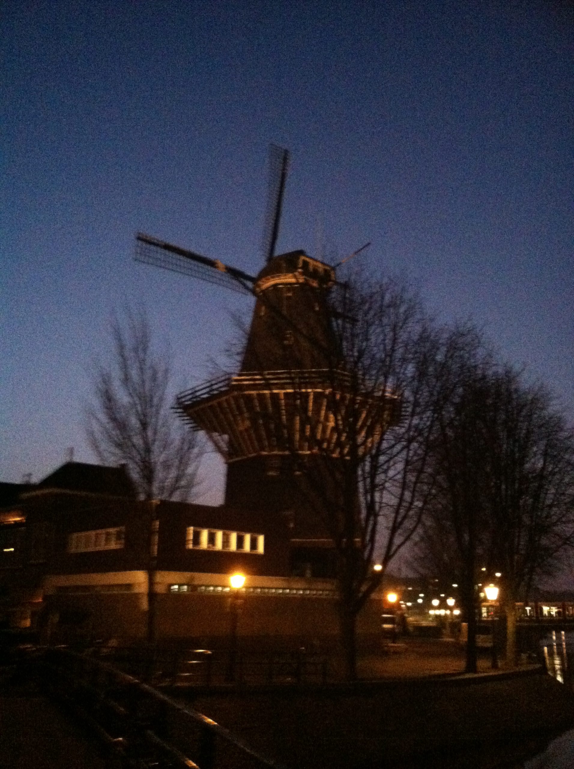 An Evening at the Windmill: The picture shows a traditional windmill during dusk. The sky is a deep blue as the sun has set. The windmill is tall and has four large blades; it appears to be made of wood. There's a balcony-like structure around the middle of the windmill. In the background, there are leafless trees and a building with lights on. The area looks like it might be a small town or a suburb. There's a chain fence in the foreground and a street lamp emitting a warm yellow light. The atmosphere seems calm and serene.