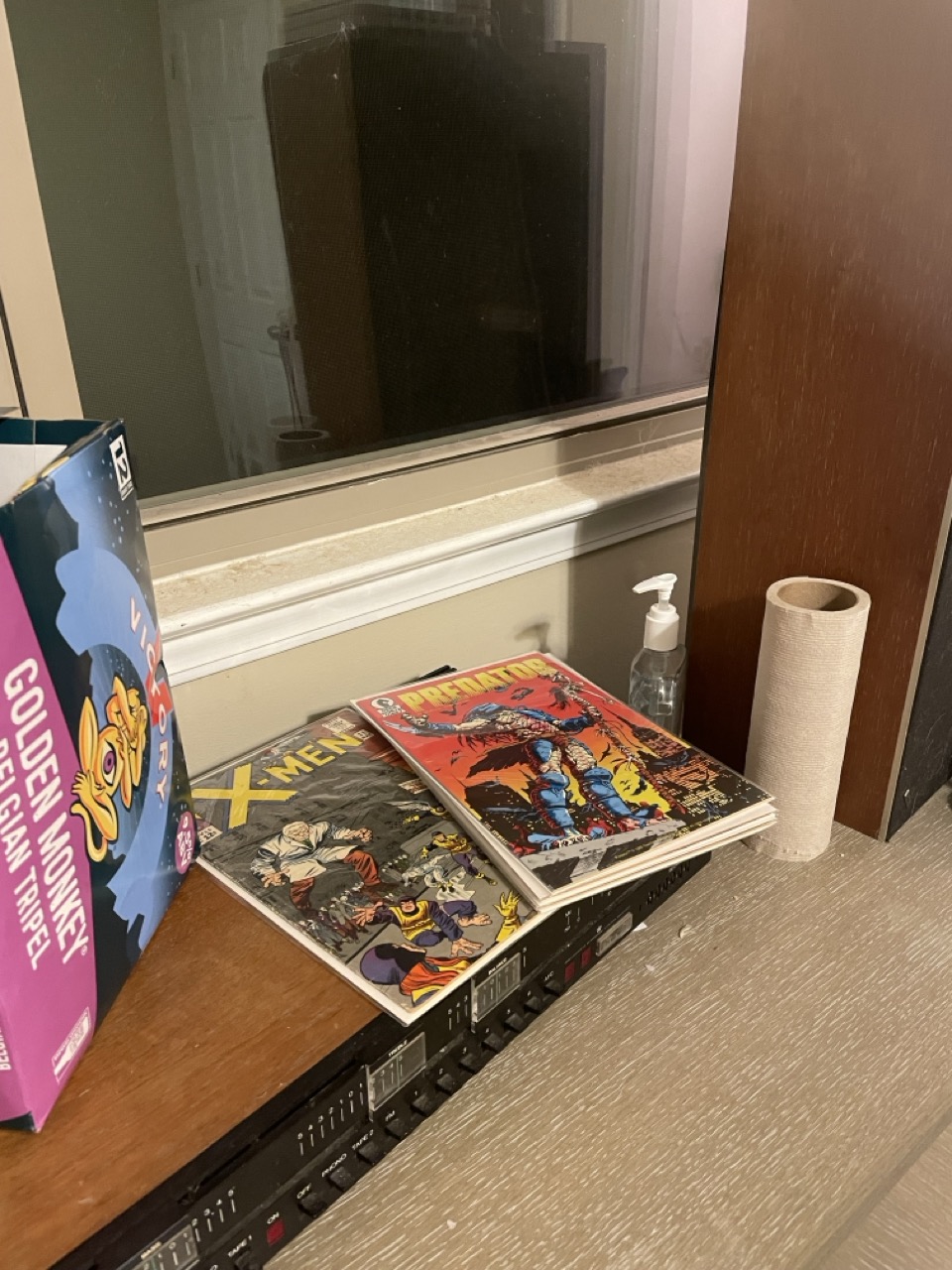 Some Belgian Beer for the Blind Nerd: The picture shows a corner of a room with a wooden ledge against a wall. On the ledge, there are several items. On the left, there is a colorful box with a cartoonish design, which seems to be a packaging for a product named "Golden Belgian Monkey Triple". Next to it, there are three comic books with vibrant covers. The one on top is an X-Men comic, featuring characters in action poses. To the right of the comic books, there is a small bottle with a pump, possibly a hand sanitizer, and a roll of paper towels. Above the ledge, there is a large dark screen, possibly a television, mounted on the wall. The wall on the right side is brown, and the wall behind the television is painted in a lighter color. The ledge has a textured finish.