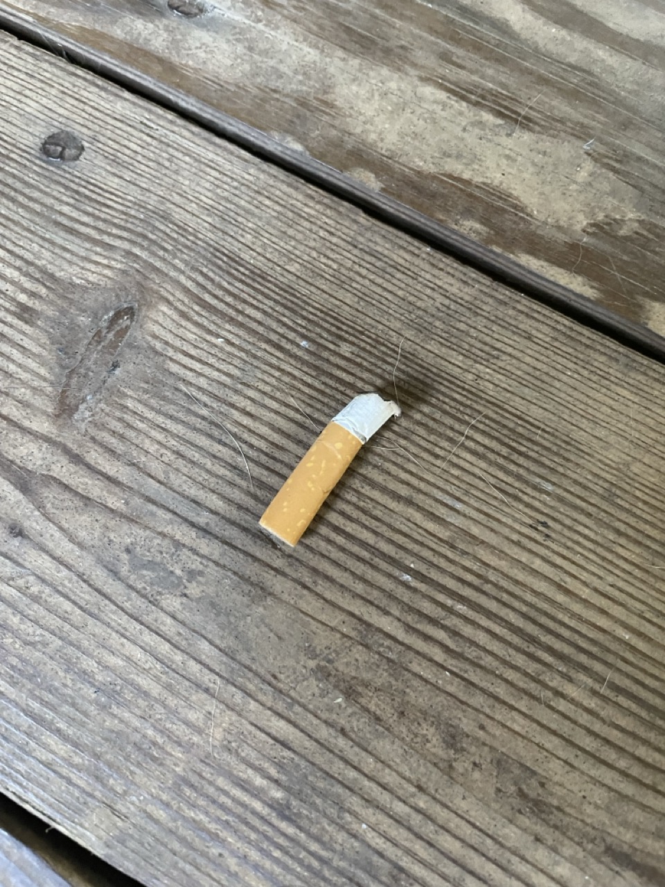 The Chain-Smoking Grandma: The picture shows a close-up of a wooden surface, possibly a floor or a table. The wood has a weathered look with visible grain patterns and a few small dark spots. There is a cigarette butt lying on the wooden surface towards the bottom right corner. The cigarette butt has an orange filter and a small portion of white paper with some ash at the end.