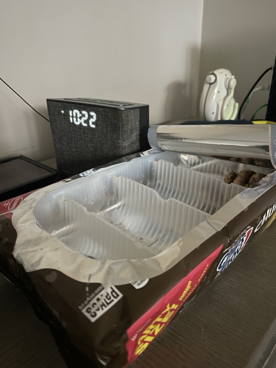 Who Ate the F#@%ing Cookies?: The picture shows a few items on a wooden surface, possibly a desk or a table. In the foreground, there is an open pack of snacks. The packaging is brown with a white plastic tray inside. The tray is almost empty, with just a few pieces of snacks left. The packaging has some text on it, including the word "SNACK" in yellow and red. In the background, there is a digital clock with a dark gray fabric exterior. The clock displays the time as 10:22. There is also a white object next to the clock, which looks like a game controller with buttons and a joystick. Additionally, there is a book or a notebook with a white cover underneath the clock. The background wall is light-colored.