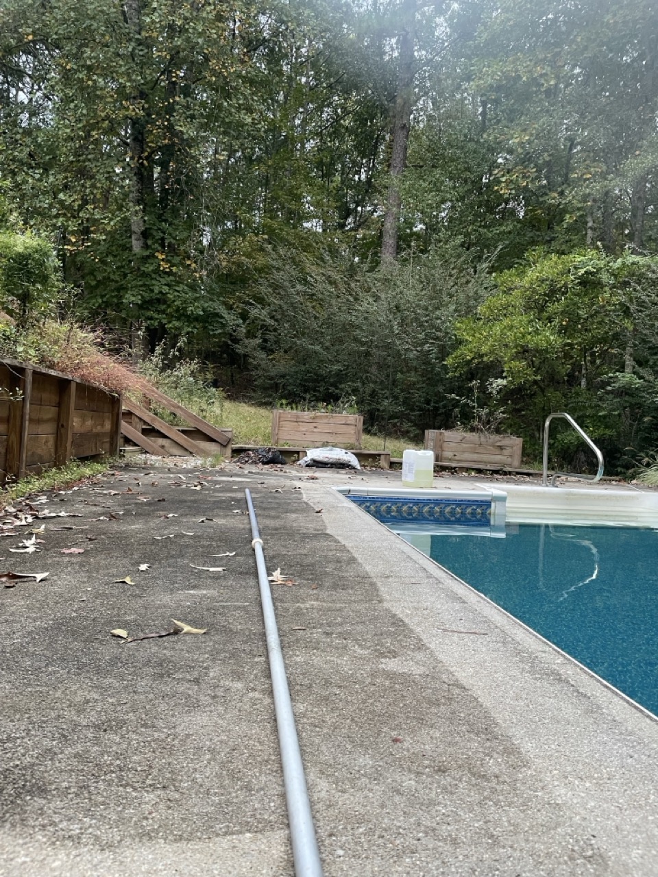 Festivus Preparations: The picture shows an outdoor setting with a swimming pool. The pool is on the right side of the image and has a blue water with a decorative blue tile border. There is a metal pole lying horizontally on the concrete ground next to the pool. The ground has some scattered leaves on it. On the left side, there is a wooden fence that is slightly tilted and behind it, there are dense trees and shrubs. The trees have green leaves with some turning yellow, indicating it might be autumn. There is also a wooden structure with what appears to be two compartments or boxes near the fence.