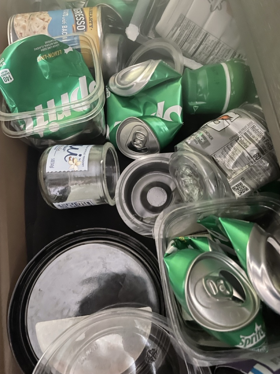 The Misplaced Recycling: The picture shows a collection of various cans and containers. There are several green cans which appear to be Sprite soda cans. There are also some transparent plastic containers with black lids. Additionally, there is a can with a label showing a picture of a chicken, which might be canned chicken. The items are all jumbled together.