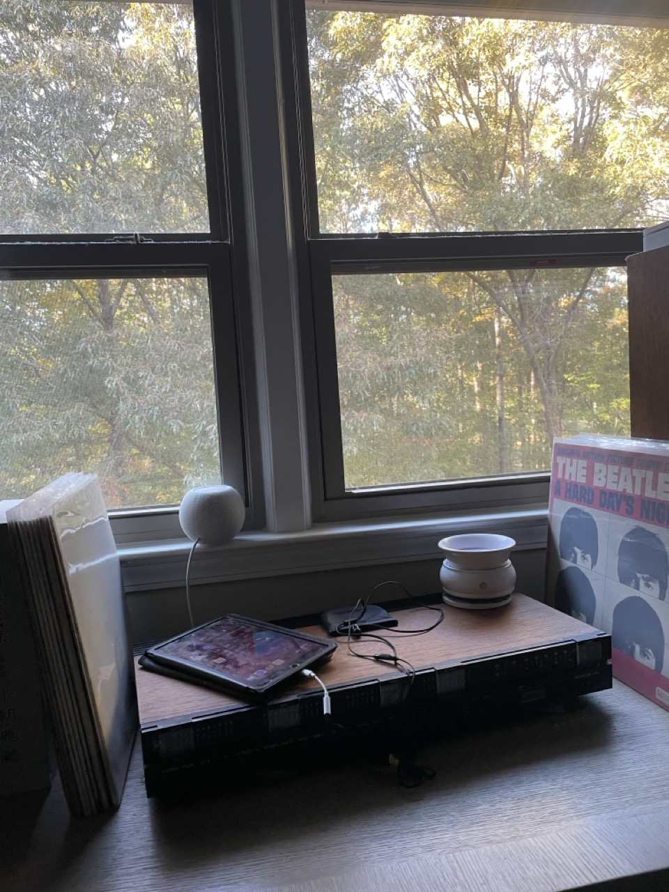 Zoom Conference Comedy Rehearsal: The picture shows a cozy indoor setting near a window. The window is large with a view of lush green trees outside, and the sunlight is filtering through the leaves. On a wooden table near the window, there is a tablet with its screen on, showing app icons. The tablet is lying on top of a black rectangular object, possibly a case or a book. Next to the tablet, there is a small white spherical speaker with a cable attached to it. On the right side of the speaker, there is a white circular object, which looks like a small container or a decorative item. On the far right, there is a vinyl record cover leaning against the wall with "The Beatles A Hard Day's Night" written on it, featuring pictures of the band members. On the left side of the tablet, there are some white papers or documents stacked vertically. The overall atmosphere of the picture is calm and serene with a touch of nostalgia due to the vinyl record.