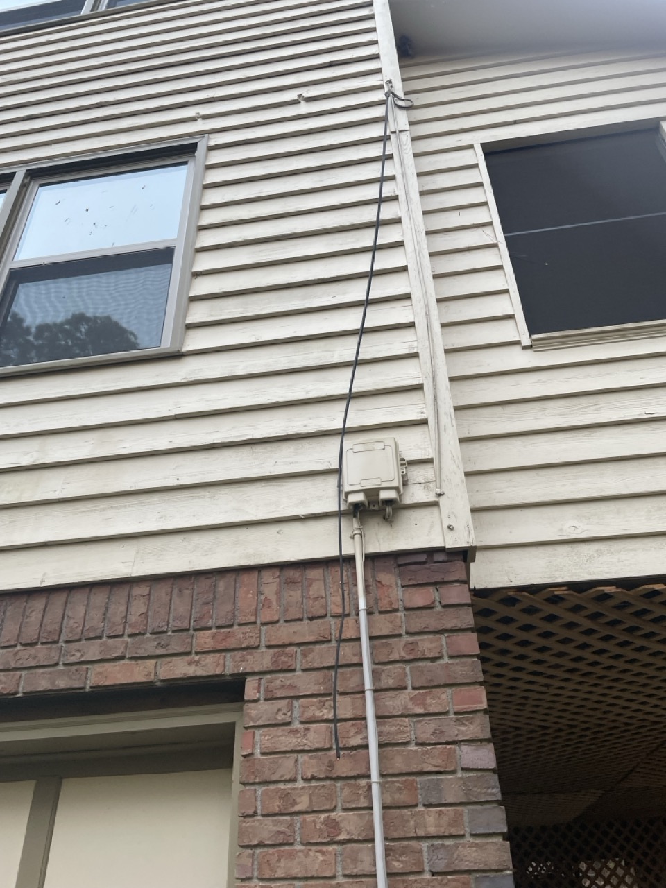 The Intermittent Internet: The picture shows the exterior of a two-story house. The house has beige horizontal siding on the upper level and a brick facade on the lower level. There is a window on each floor; the upper window is open. A white electrical box is mounted on the siding where the brick and siding meet, with a conduit running vertically up the wall and a cable running down. There is also a lattice structure at the bottom right corner of the image.