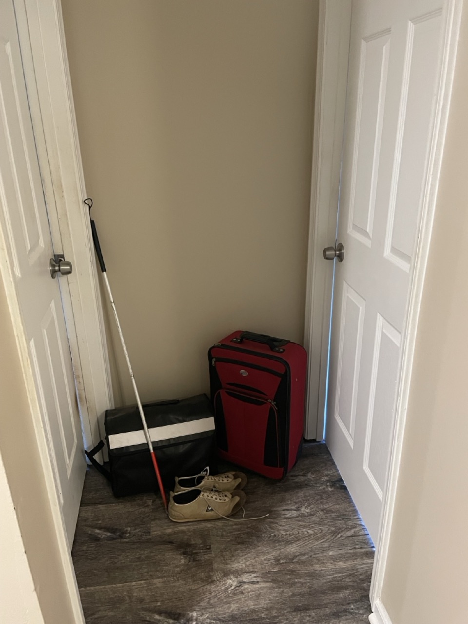 Today Is Election Day: The picture shows a small hallway or alcove in a house. The walls are painted in a light beige color and the floor has dark wood planks. On the left side, there is a white door with a silver handle, slightly ajar. Leaning against the wall next to the door is a white cane with a red bottom and a black handle. On the right side, there is another white door which is closed, with a silver handle. In the corner between the two doors, there are a few items placed on the floor: - A large red suitcase with black trim and a handle at the top. - A black rectangular bag with a white stripe in the middle. - A pair of beige shoes with white laces. The space looks like it might be used for storage or as a place to keep items that are frequently used when leaving the house.